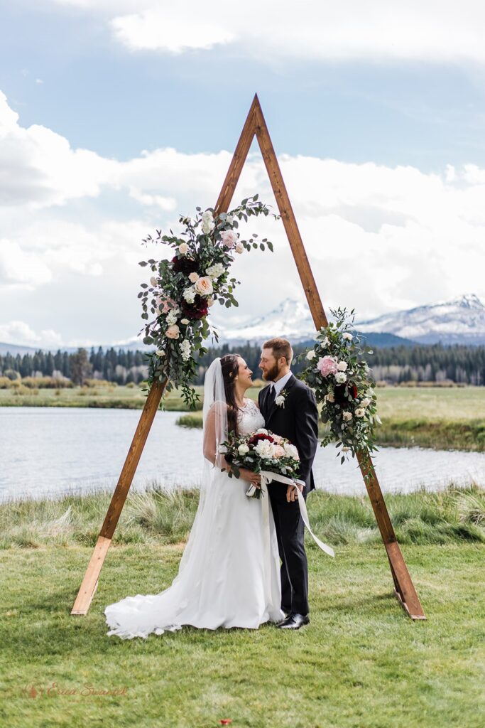 A bride and groom admire one another beneath an a-frame arch decorated with rose floral arrangements near a lake and mountains at Black Butte Ranch. 