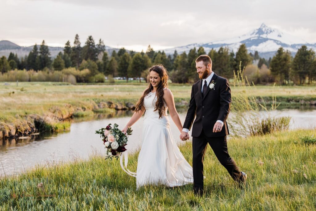 A couple dressed in formal wedding attire hold hands while walking along a river bank during their wedding at Black Butte Ranch. 