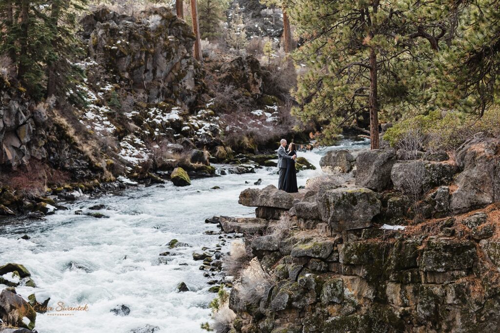 A couple dressed in dark, formal wedding attire dances on a cliff's edge during their Dillon Falls elopement in Oregon. 
