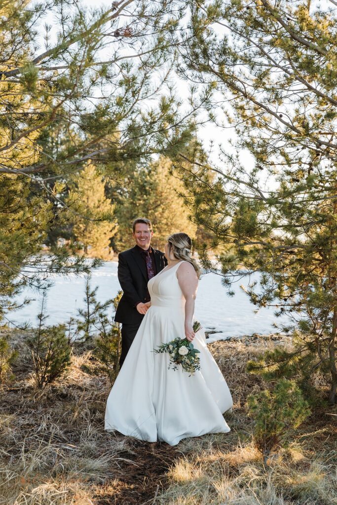 A man wearing a black suit holds his bride's hand who is wearing a wedding gown and holding a white rose bridal bouquet near Sunriver. 