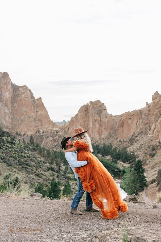 A bride in a orange boho dress and orange cowboy hat is lifted up by her groom during their Smith Rock State Park elopement. 