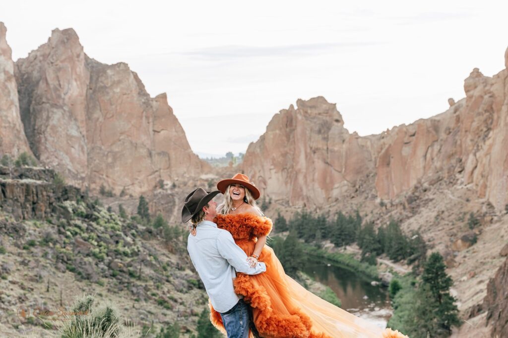A bride in a orange boho dress and orange cowboy hat is lifted up by her groom who is wearing a cowboy hat, western shirt and jeans during their Smith Rock State Park elopement. 