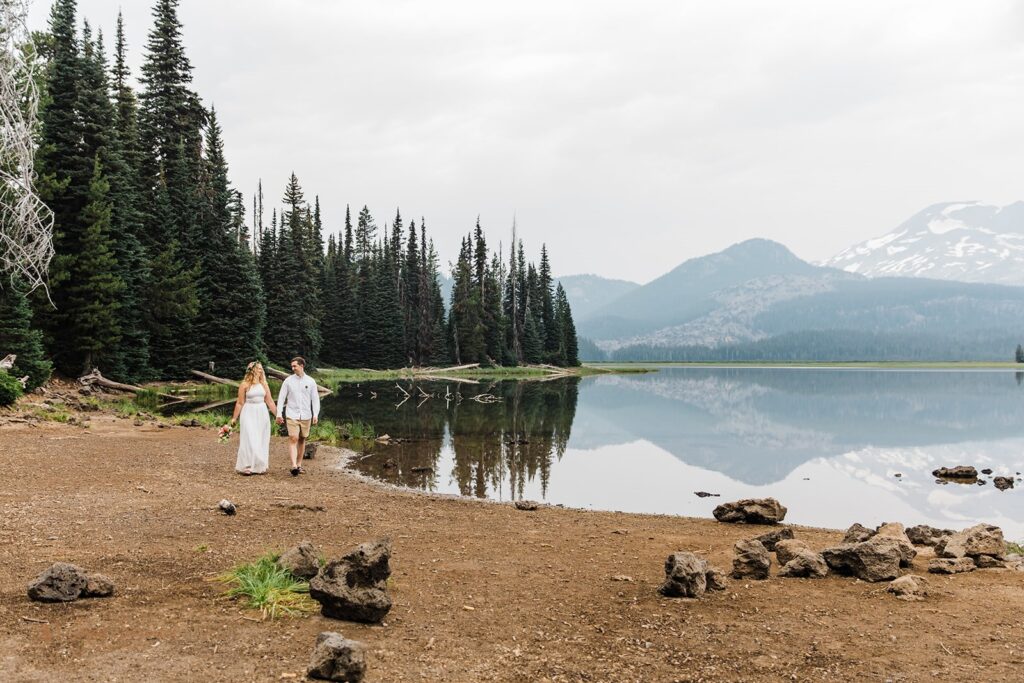 An Oregon elopement couple holds hands while walking along the shores of Sparks Lake, an intimate Bend wedding location.