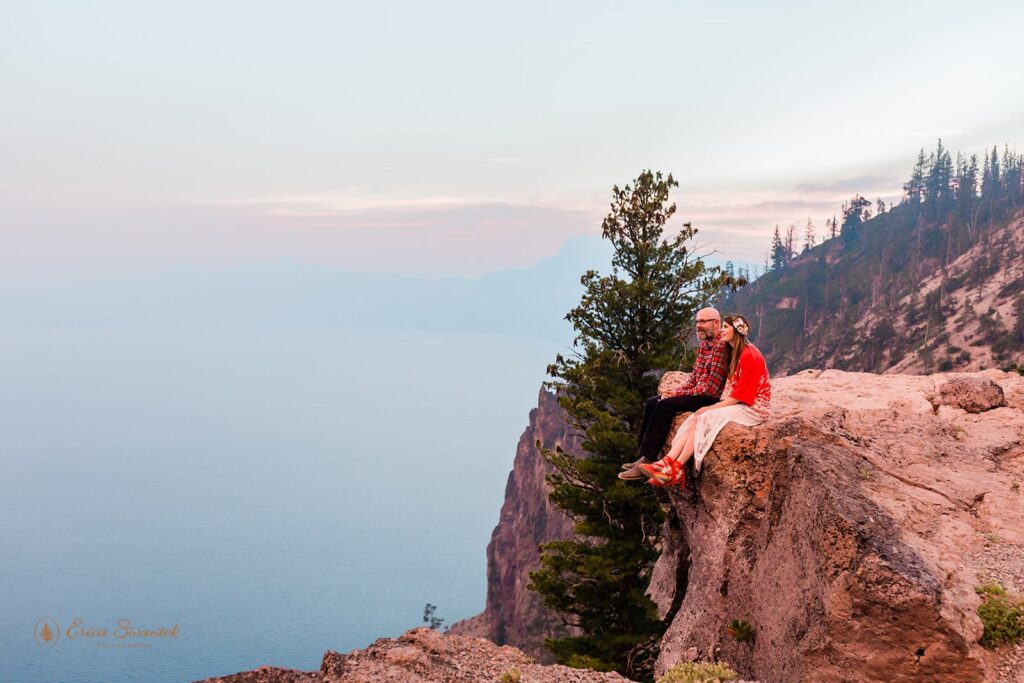 An adventure elopement couple sit on a cliff's edge overlooking Crater Lake.