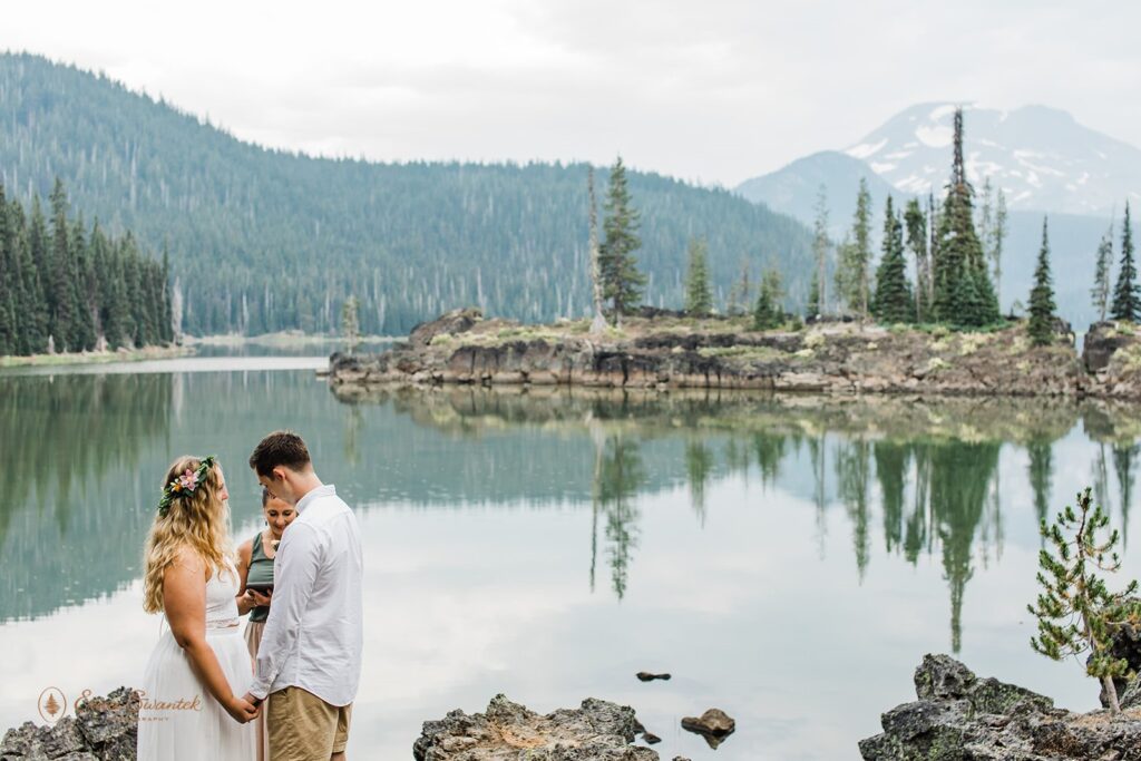An intimate wedding couple recites vows with their Oregon officiant on the shores of Sparks Lake near Bend. 