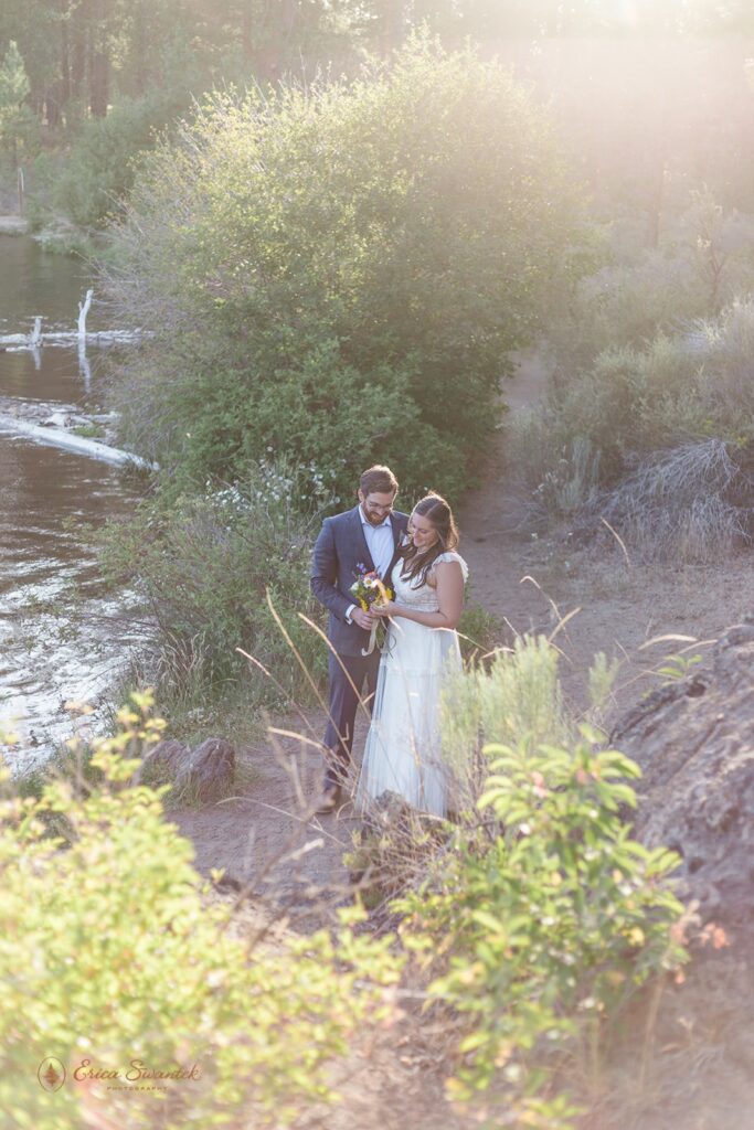 A Bend elopement couple in formal wedding attire embraces along the banks of the Deschutes River. 