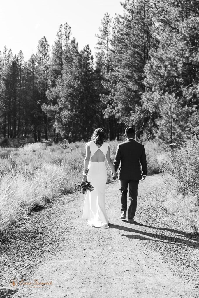 A hiking elopement couple in formal wedding attire walks along the Deschutes River Trail while holding hands. 