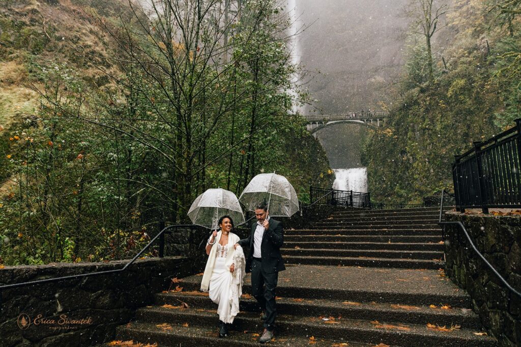 Bride and groom walking up steps in the rain holding clear umbrellas with a stunning waterfall in the background.