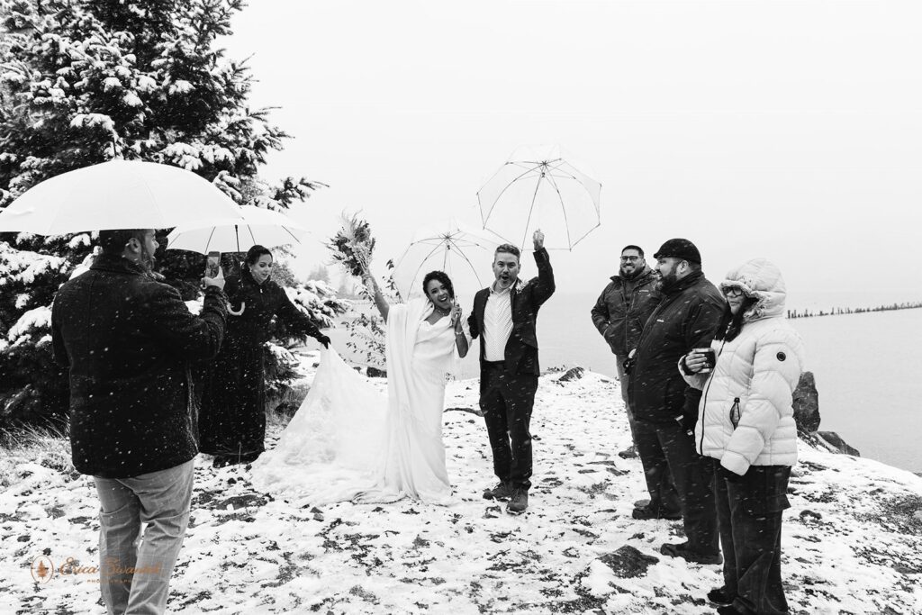 Bride and groom celebrate under clear umbrellas with their guests surrounding them on a snow-covered cliffside.