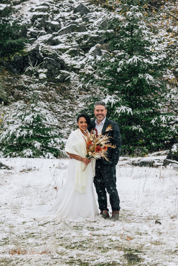 Bride and groom share an intimate moment, standing close together with snow falling and a backdrop of snow-covered trees.