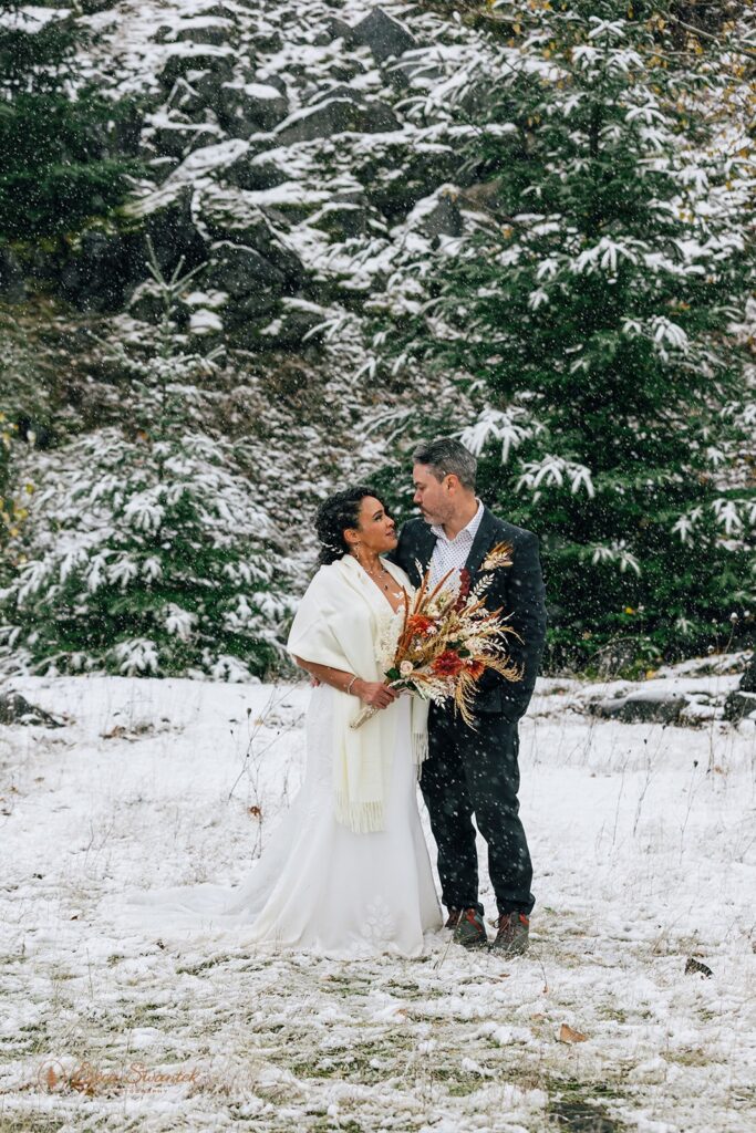 Bride and groom share an intimate moment, standing close together with snow falling and a backdrop of snow-covered trees.