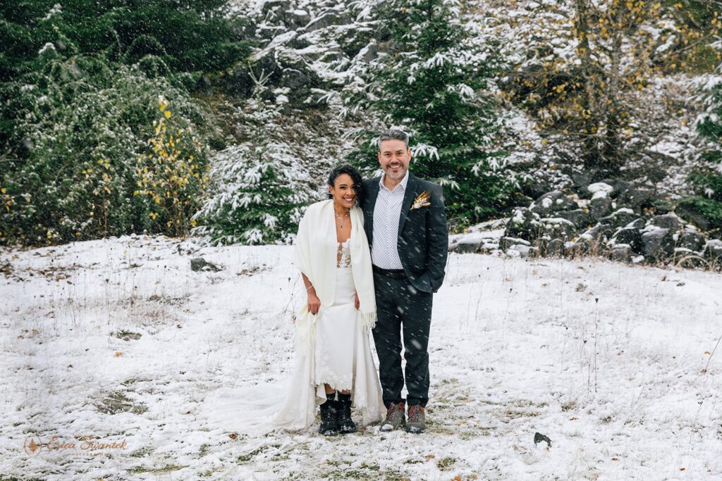 Bride and groom share an intimate moment, standing close together with snow falling and a backdrop of snow-covered trees.