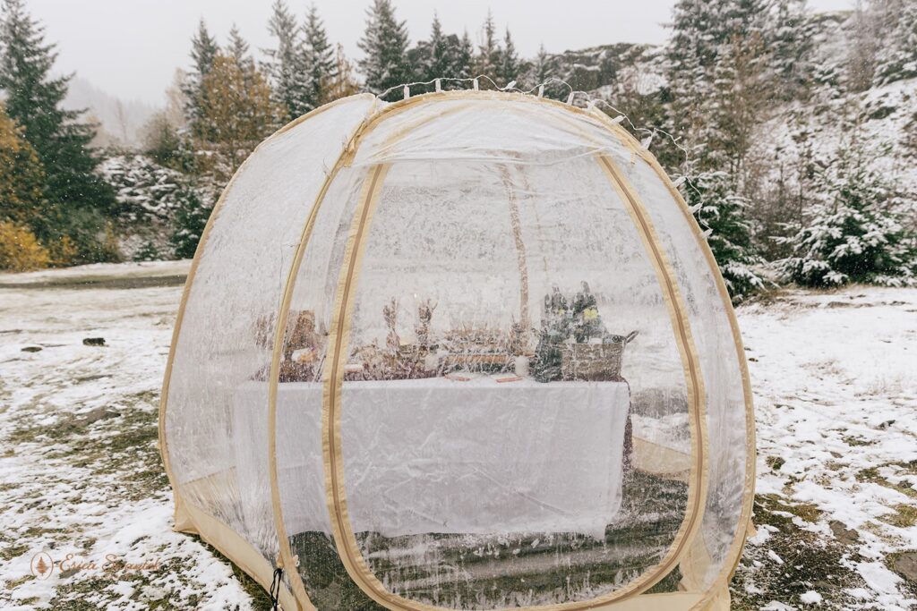 Clear tent set up for a winter elopement, with a snow-covered landscape in the background, featuring a decorated table inside.