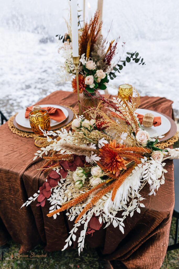 A beautifully decorated table set for two with an earthy-toned tablecloth, dried floral centerpiece, and gold accents inside a clear tent with a snowy background, perfect for a boho style