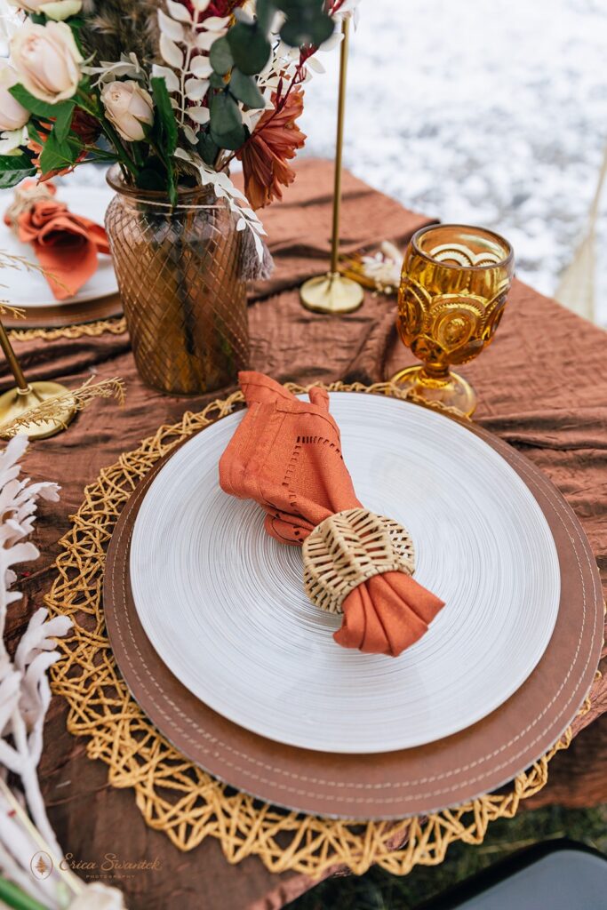 A beautifully decorated table set for two with an earthy-toned tablecloth, dried floral centerpiece, and gold accents inside a clear tent with a snowy background, perfect for a boho style