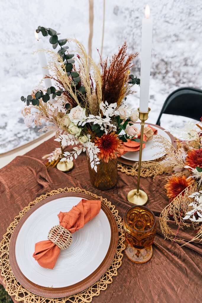 A beautifully decorated table set for two with an earthy-toned tablecloth, dried floral centerpiece, and gold accents inside a clear tent with a snowy background, perfect for a boho style