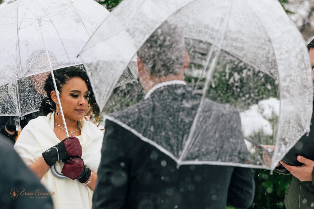 Bride and groom standing close under clear umbrellas during a light snow shower. The bride is wrapped in a white shawl and wearing maroon gloves.