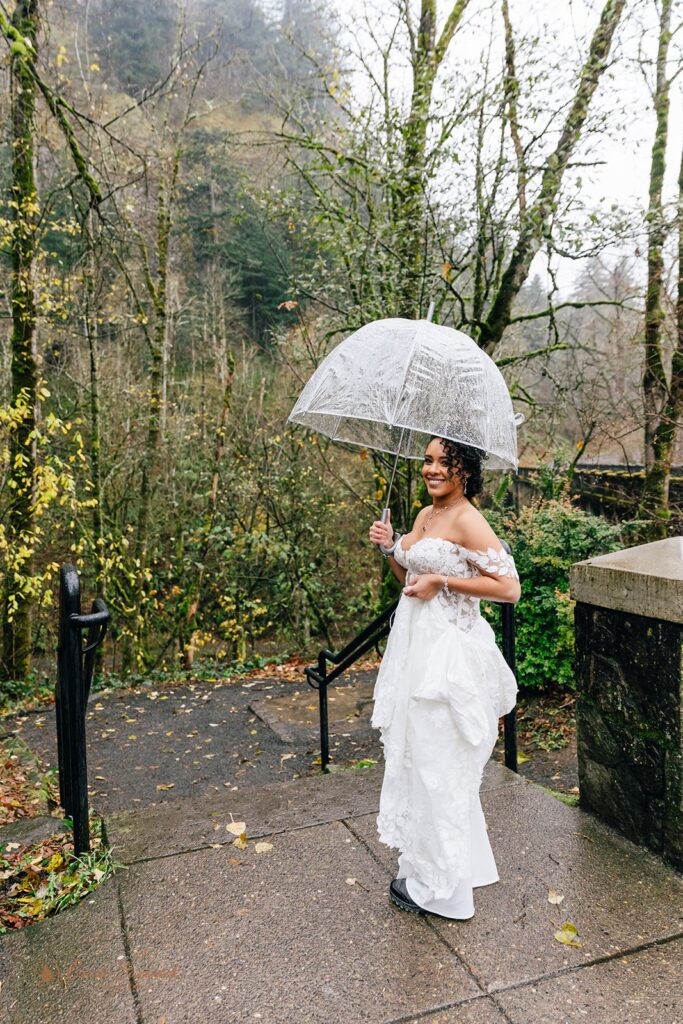 A bride walking down stone steps, holding a clear umbrella and the train of her white wedding dress. 