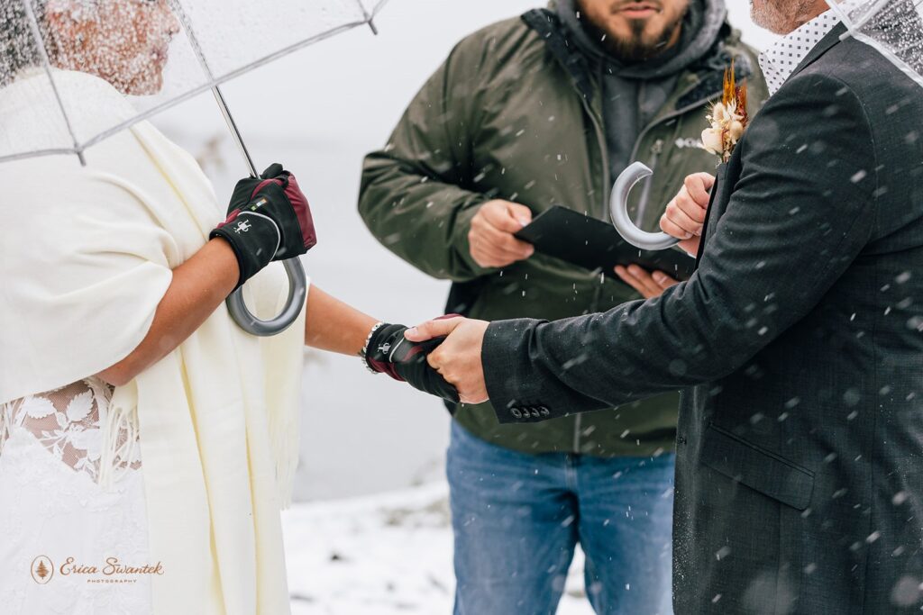 Bride and groom standing close under clear umbrellas during a light snow shower. The bride is wrapped in a white shawl and wearing maroon gloves.