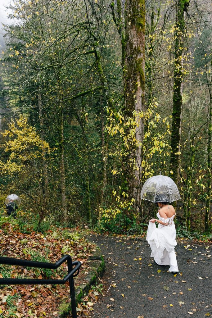 A bride walking down stone steps, holding a clear umbrella and the train of her white wedding dress. 