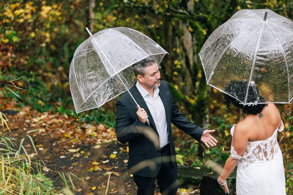 bride and groom elopement first look under clear umbrellas in a forested area