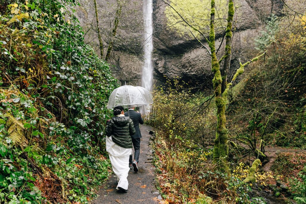 bride and groom under clear umbrellas walking towards a waterfall in columbia river gorge