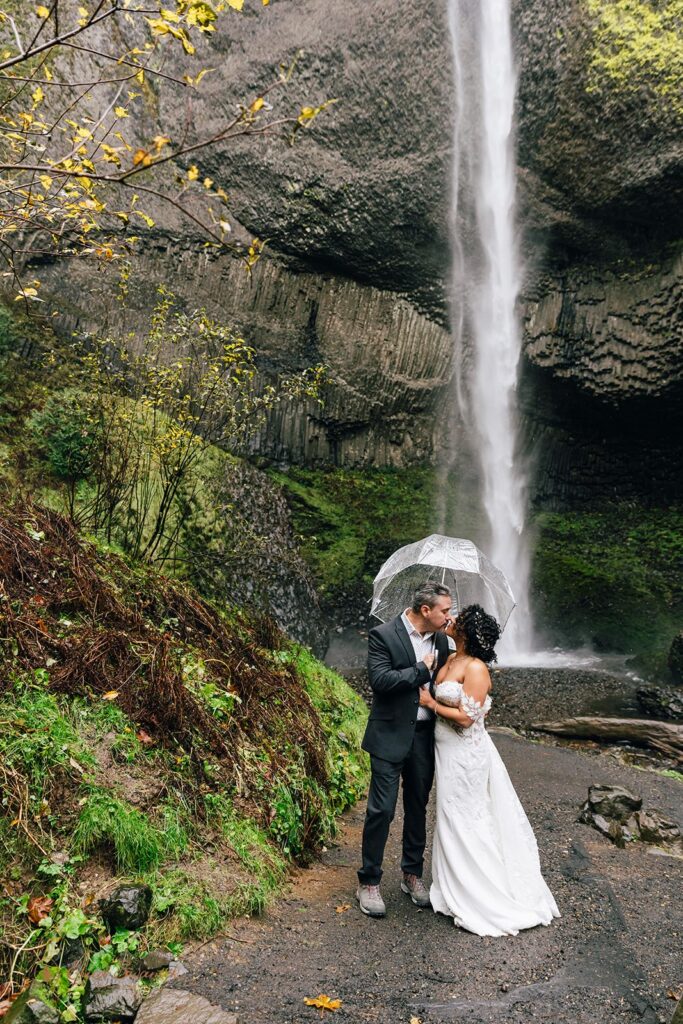 A couple kissing under a clear umbrella in front of a waterfall during their Columbia River Gorge elopement day, surrounded by green moss and rocky terrain.