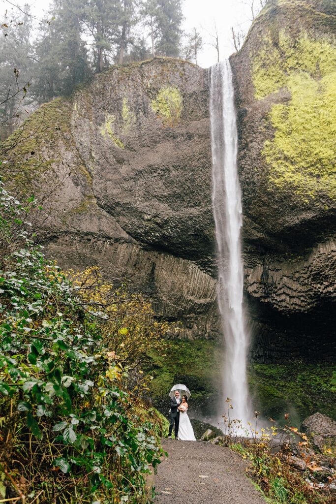 A bride and groom standing in front of a large waterfall, both holding clear umbrellas. The waterfall cascades down a mossy cliff.