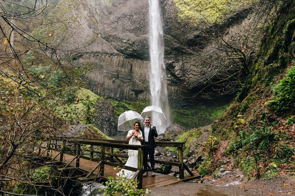 A bride and groom standing in front of a large waterfall, holding clear umbrellas. The bride is in a white wedding dress, and the groom is in a dark suit. The scene is surrounded by lush greenery and mist from the waterfall.