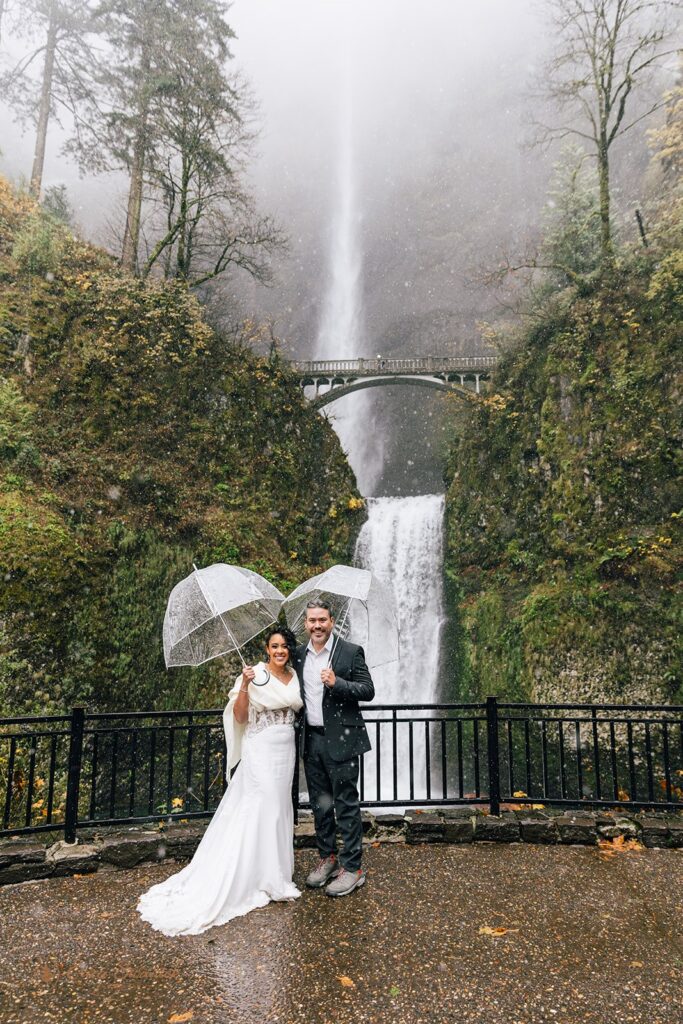 A couple posing under a clear umbrella in front of a waterfall during their Columbia River Gorge elopement day, surrounded by green moss and rocky terrain.