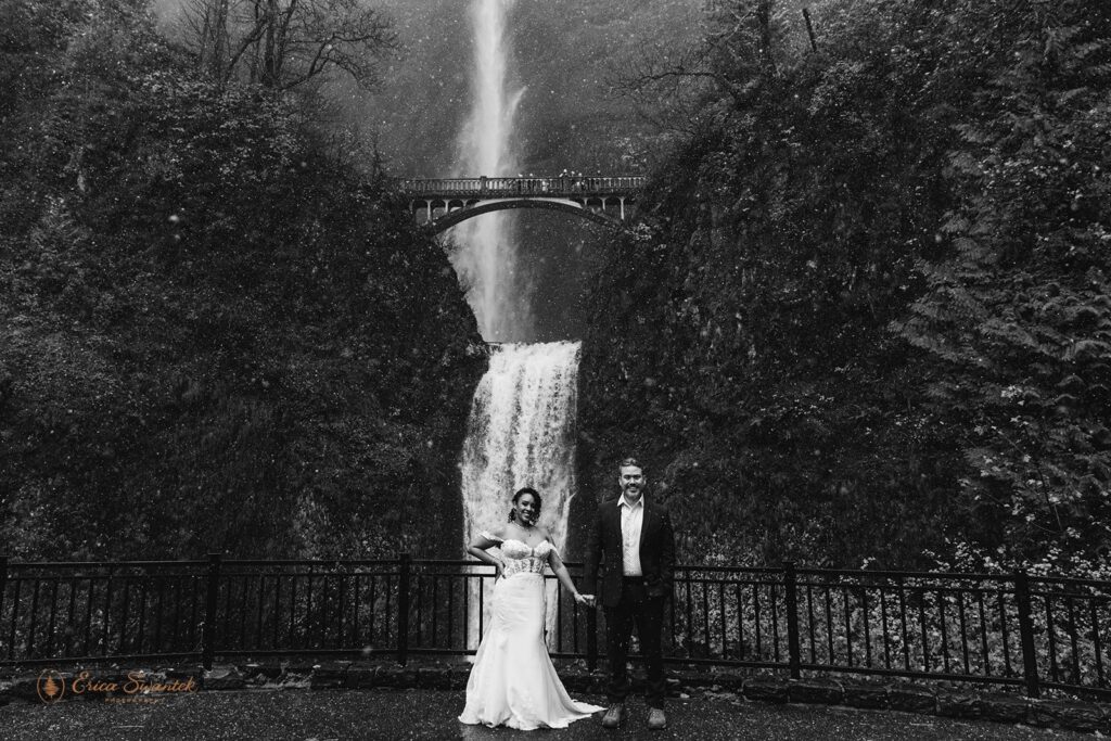 A bride and groom standing in front of a large waterfall, both holding clear umbrellas. The bride is in a white wedding dress, and the groom is in a dark suit. The waterfall cascades down a mossy cliff with a bridge in the background.