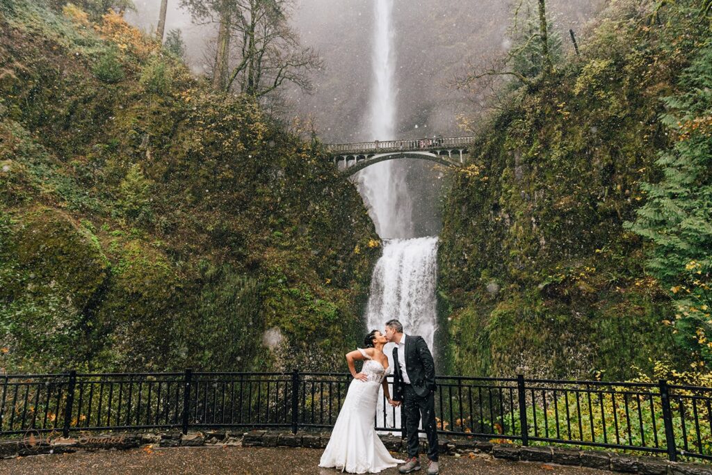 A bride and groom standing in front of a large waterfall, both holding clear umbrellas. The bride is in a white wedding dress, and the groom is in a dark suit. The waterfall cascades down a mossy cliff with a bridge in the background.