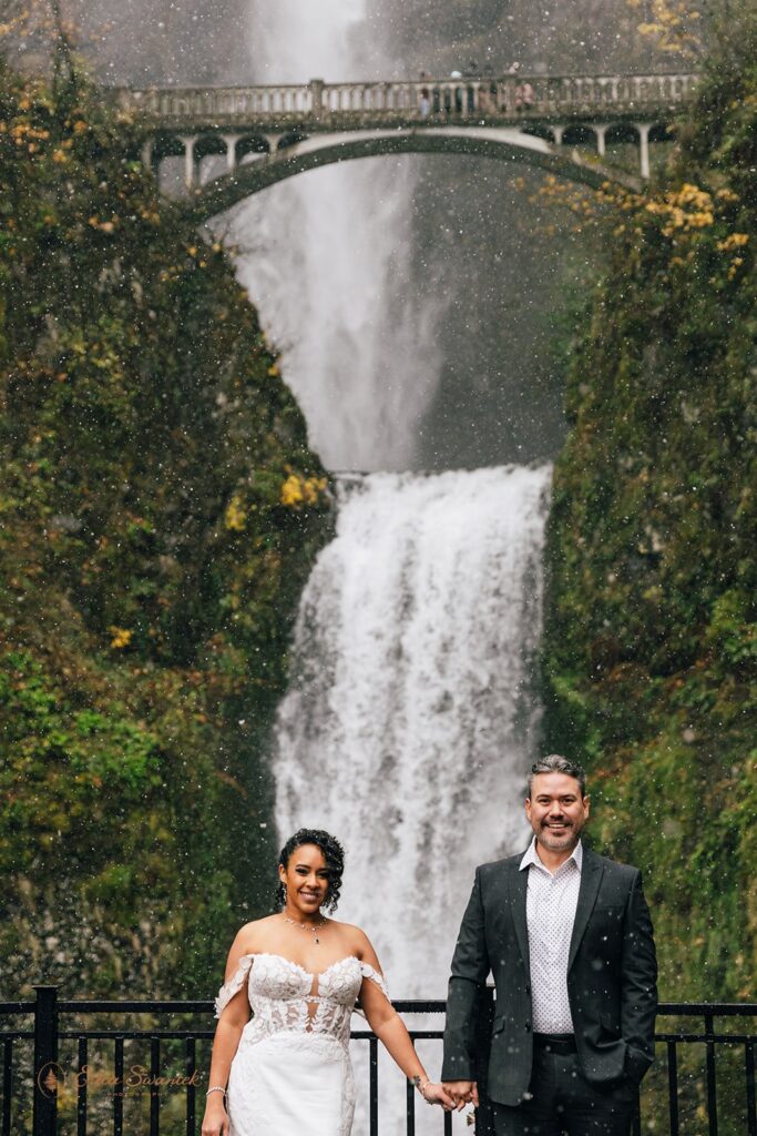 A bride and groom standing in front of a large waterfall, both holding clear umbrellas. The bride is in a white wedding dress, and the groom is in a dark suit. The waterfall cascades down a mossy cliff with a bridge in the background.