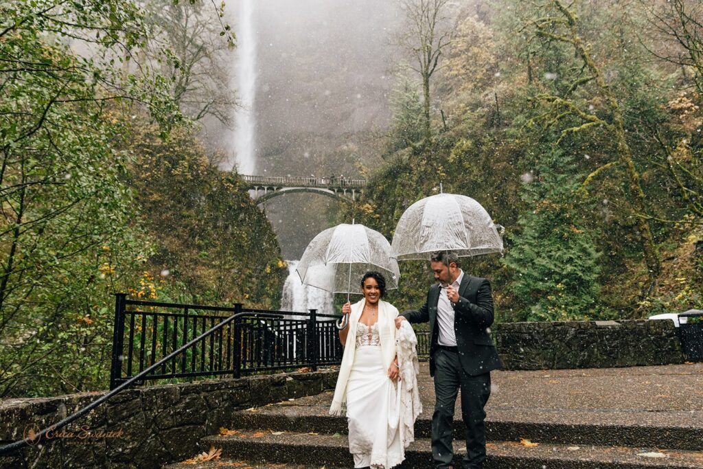 A couple walking down stairs in front of a large waterfall, each holding a clear umbrella. The bride is wearing a white wedding dress, and the groom is in a dark suit. The waterfall and surrounding area are lush with greenery.