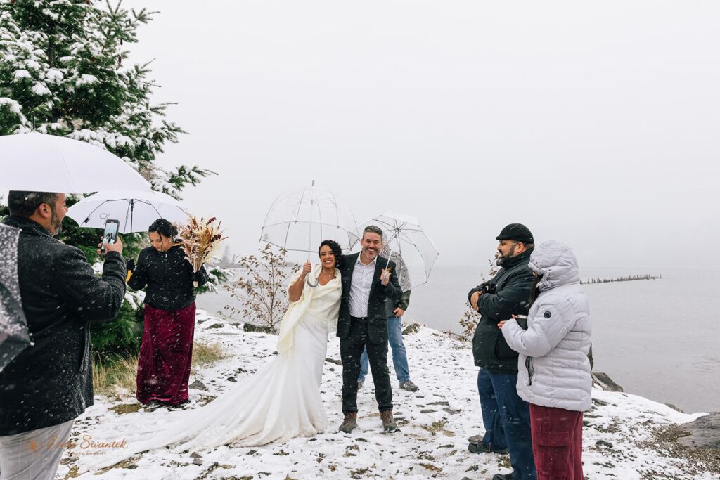 Bride and groom celebrate under clear umbrellas with their guests surrounding them on a snow-covered cliffside.