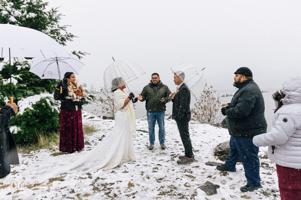 Bride and groom standing close under clear umbrellas during a light snow shower. The bride is wrapped in a white shawl and wearing maroon gloves.