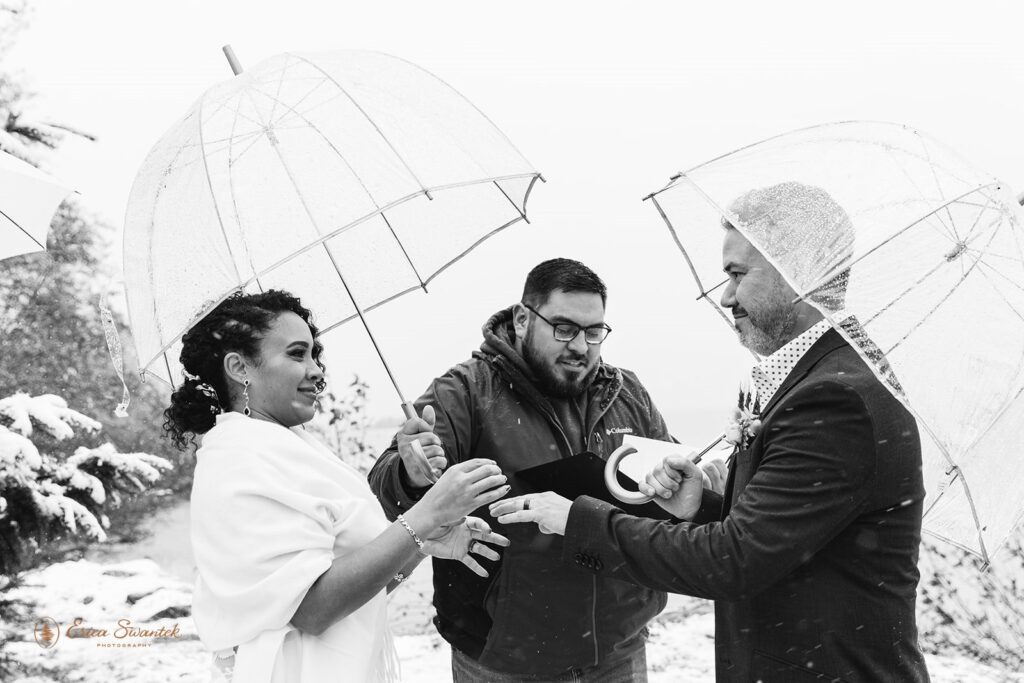 Black and white photo of the bride and groom exchanging rings under clear umbrellas while an officiant looks on during their snowy outdoor ceremony.