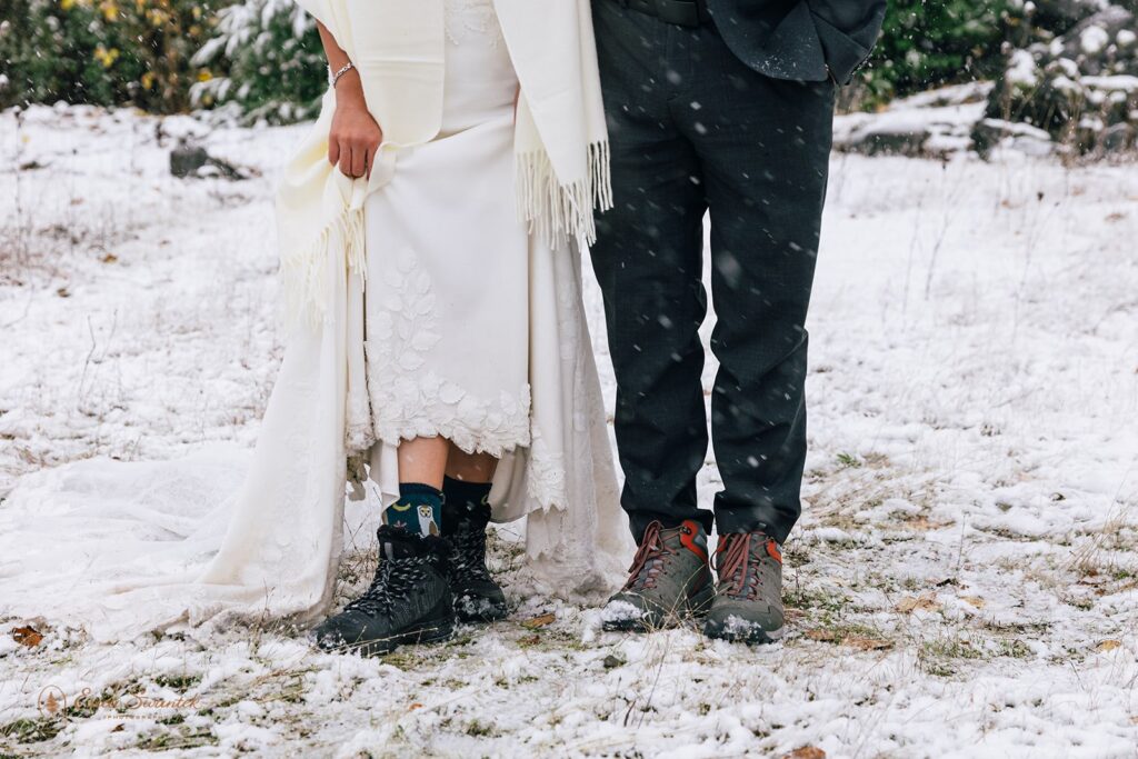 Bride and groom show off their hiking boots and cozy socks peeking out from under her wedding dress and his suit pants in the snowy landscape.