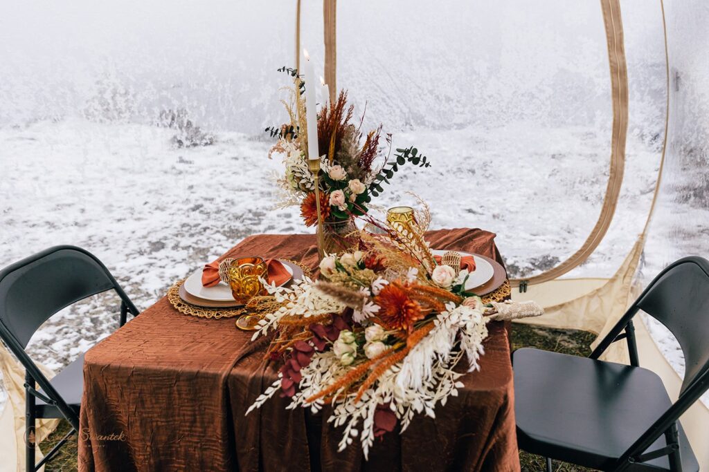 A beautifully decorated table set for two with an earthy-toned tablecloth, dried floral centerpiece, and gold accents inside a clear tent with a snowy background.