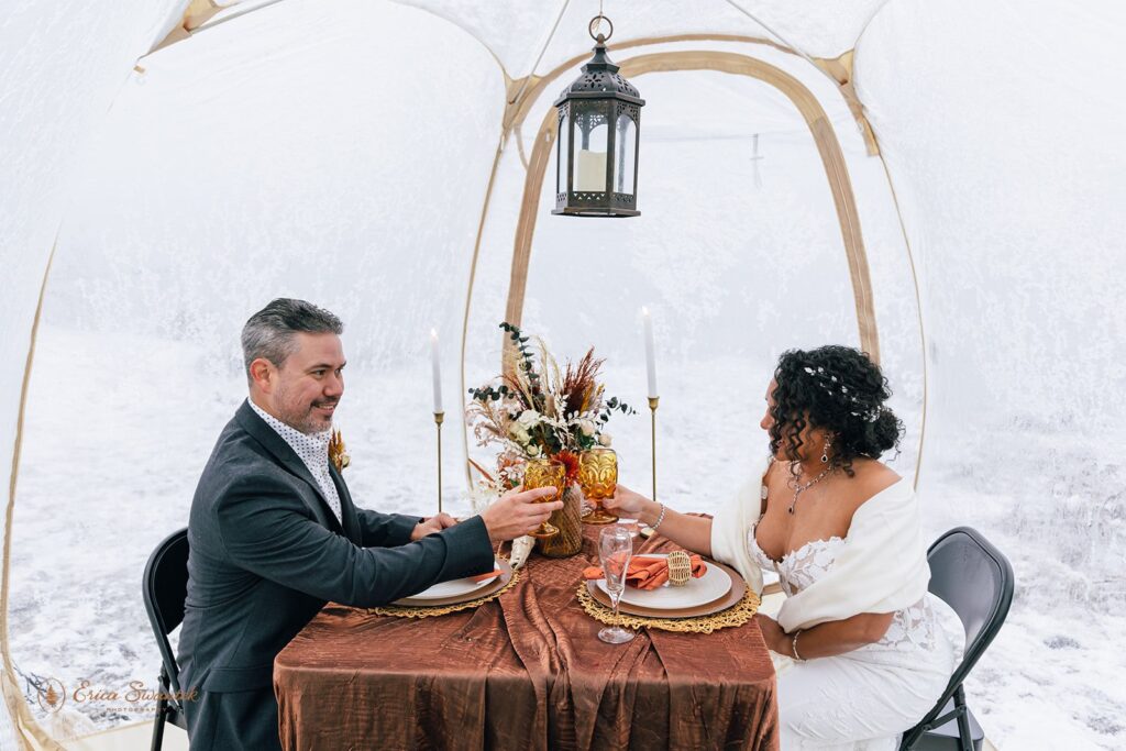 A bride and groom sitting at a decorated table inside a clear dome tent in a snowy environment. The table is set with a floral arrangement, candles, and amber goblets. The couple is toasting with their glasses, smiling at each other.