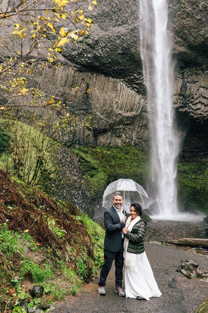 A couple kissing under a clear umbrella in front of a waterfall. The bride is wearing a white wedding dress and a green jacket, and the groom is wearing a dark suit. The waterfall is surrounded by green moss and rocky terrain.