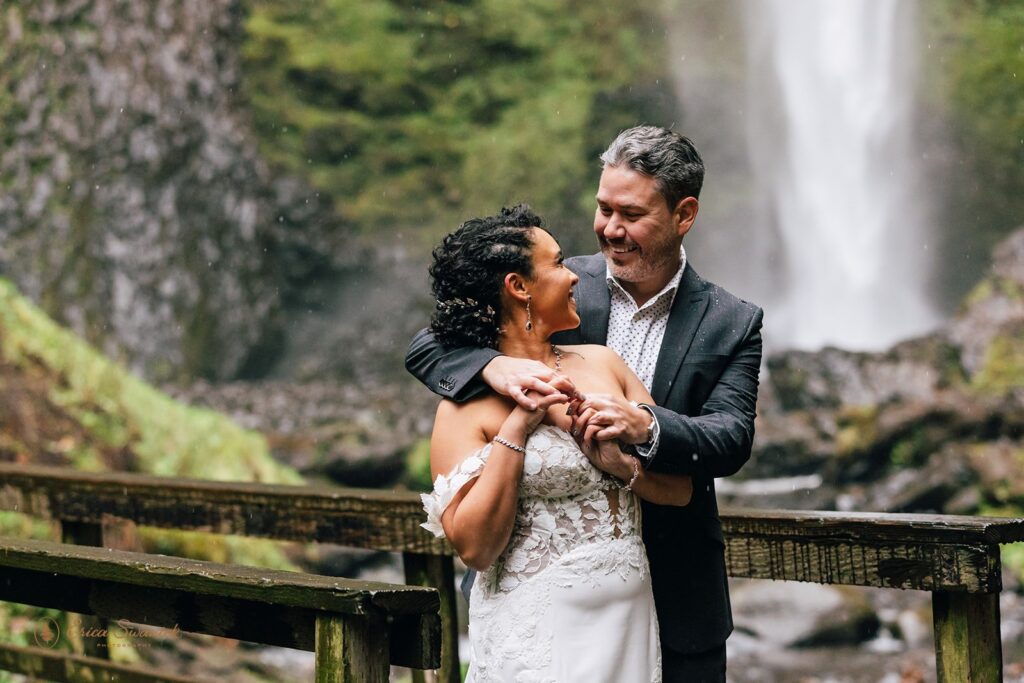 A bride and groom posing in front of a large waterfall. The bride is in a white wedding dress, and the groom is in a dark suit. The scene is surrounded by lush greenery and mist from the waterfall.