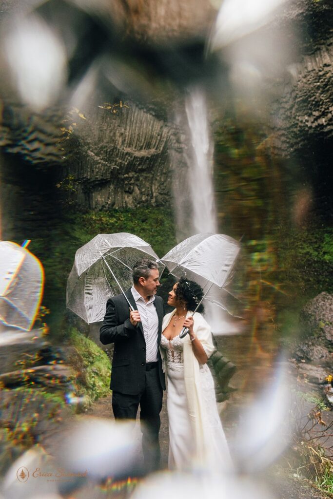 A couple kissing under a clear umbrella in front of a waterfall. The bride is wearing a white wedding dress and a green jacket, and the groom is wearing a dark suit. The waterfall is surrounded by green moss and rocky terrain.