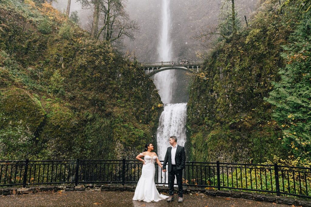 A bride and groom posing in front of a large waterfall. The bride is in a white wedding dress, and the groom is in a dark suit. The scene is surrounded by lush greenery and mist from the waterfall.