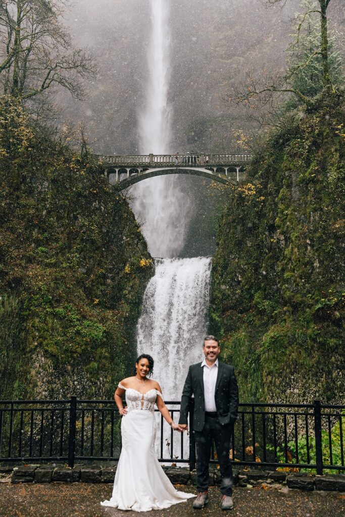 A bride and groom posing in front of a large waterfall. The bride is in a white wedding dress, and the groom is in a dark suit. The scene is surrounded by lush greenery and mist from the waterfall.