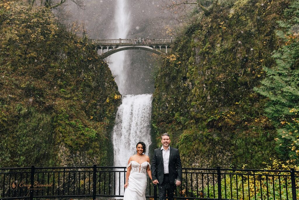 A bride and groom kissing in front of a large waterfall, holding clear umbrellas. The bride is in a white wedding dress, and the groom is in a dark suit. The scene is surrounded by lush greenery and mist from the waterfall