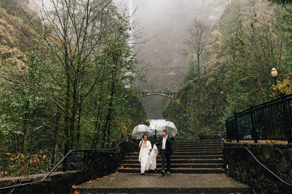 Bride and groom walking up steps in the rain holding clear umbrellas with a stunning waterfall in the background.