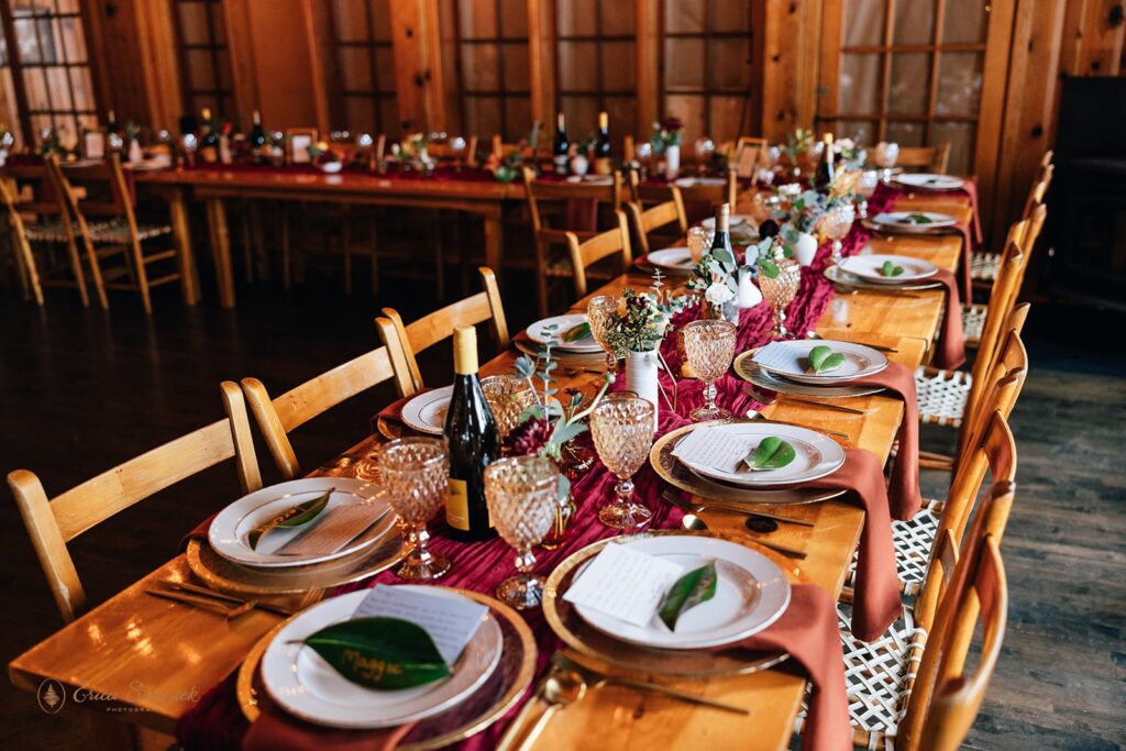 Elegant wedding reception table setup inside a wooden cabin, featuring glassware, greenery, and a rich burgundy table runner.