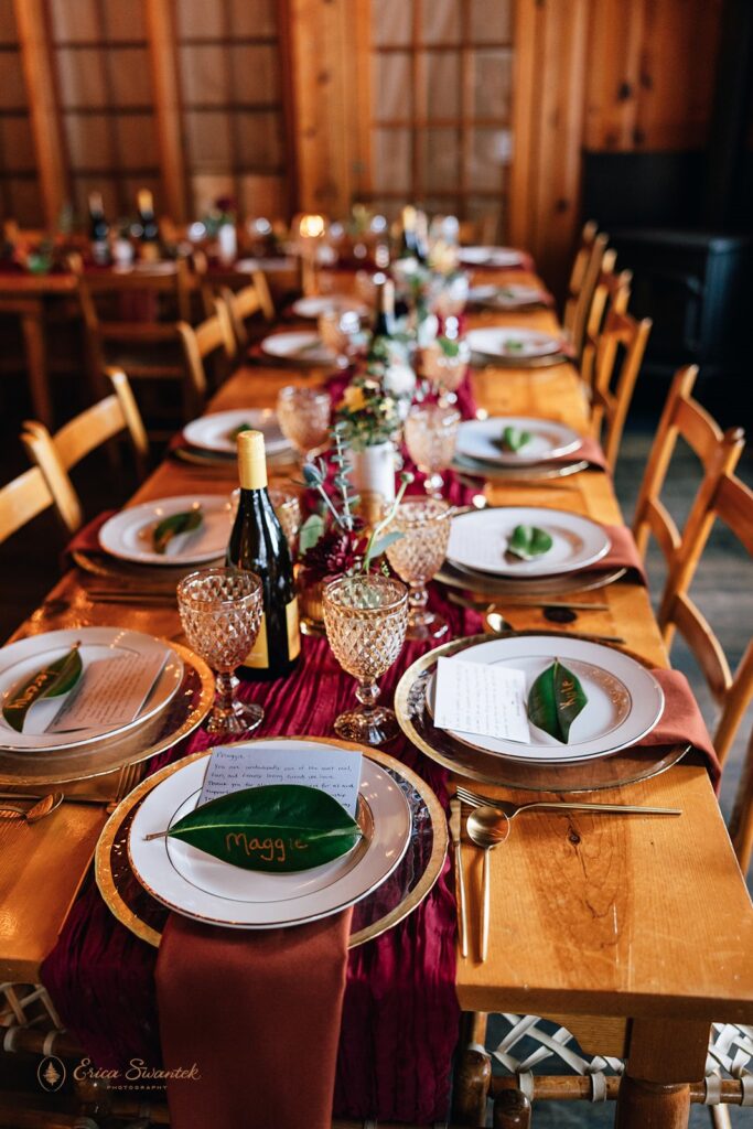 Elegant wedding reception table setup inside a wooden cabin, featuring glassware, greenery, and a rich burgundy table runner.