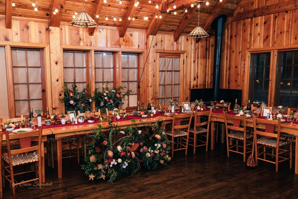 Elegant wedding reception table setup inside a wooden cabin, featuring glassware, greenery, and a rich burgundy table runner.